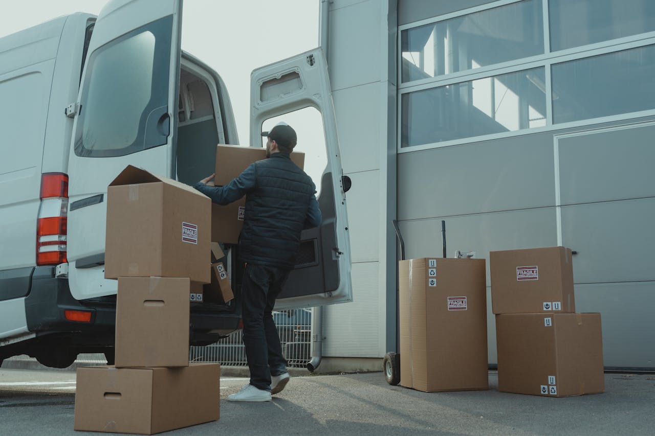 about-us A delivery man unloading cardboard boxes from a van at a warehouse during the day.