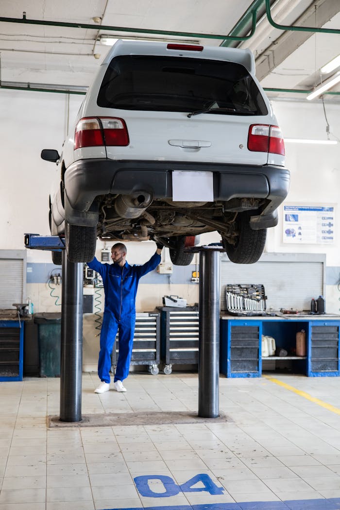 heros-img Mechanic in blue coverall inspecting a lifted car in an auto repair shop.